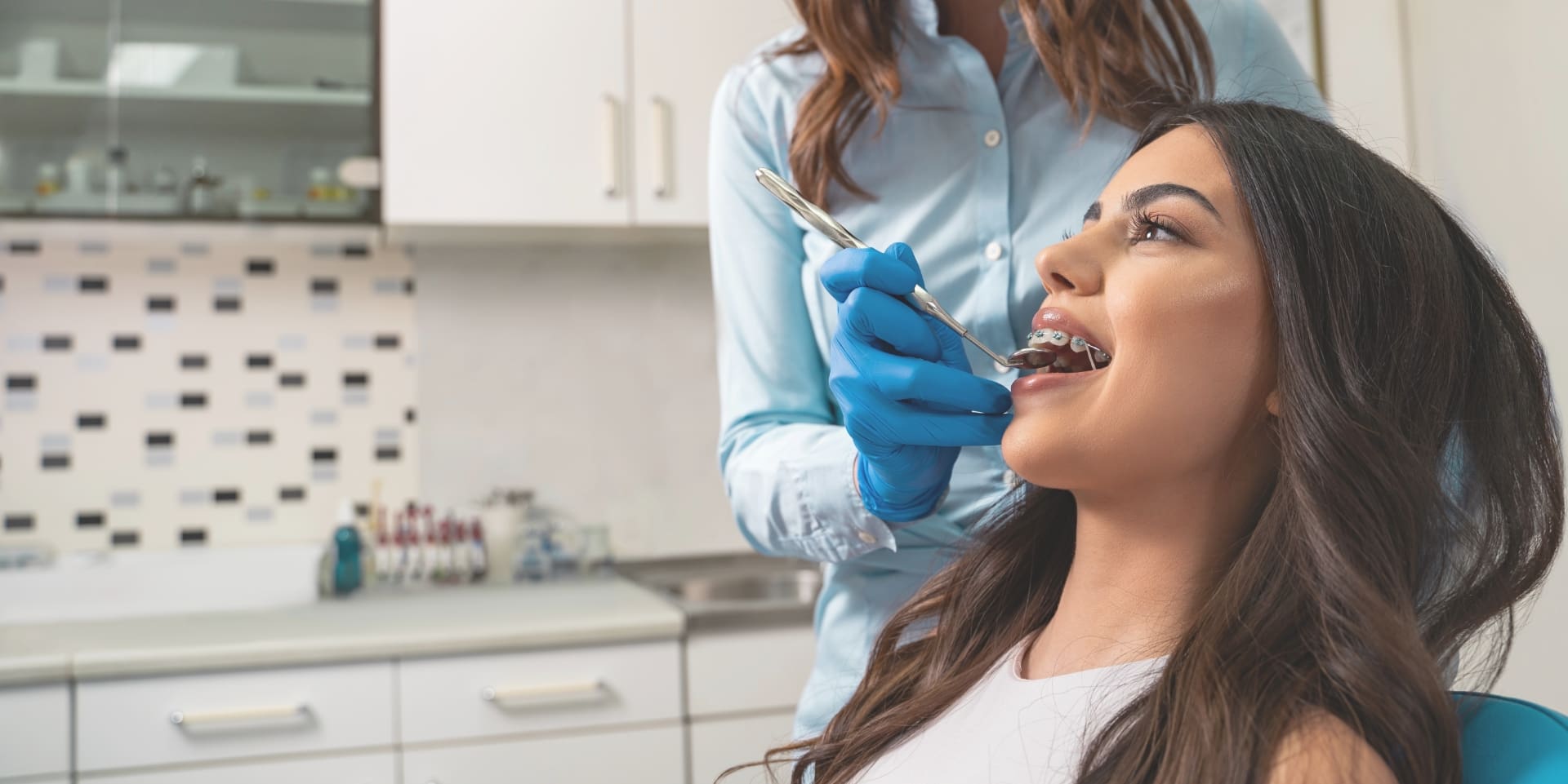 A woman sits in a dental chair while a dentist examines her teeth with tools.